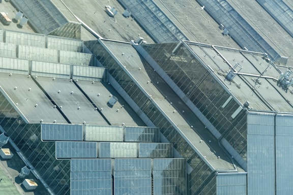 An aerial view of a large, industrial complex featuring multiple interconnected glass structures, likely greenhouses. The geometric shapes are formed by the panes of glass, and there are visible metal frames and various ventilation systems mounted on the rooftops.