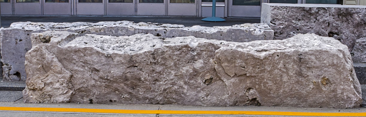 Large, rugged stone blocks are placed on a sidewalk near the entrance of a building, marked by a yellow line along the pavement.