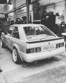 A vintage car is parked inside a workshop or garage with industrial lighting and various equipment visible in the background. The car has a boxy design common in older models, and there is a prominent exhaust pipe.