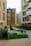 A person is sitting on a bench in a courtyard surrounded by modern apartment buildings. The architectural style consists of brick facades and large windows. There is lush green grass and several shrubs in the area. The environment appears peaceful and well-maintained.