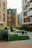 A person is sitting on a bench in a courtyard surrounded by modern apartment buildings. The architectural style consists of brick facades and large windows. There is lush green grass and several shrubs in the area. The environment appears peaceful and well-maintained.