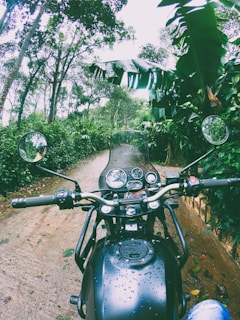 A motorcycle is parked on a dirt path surrounded by lush green foliage and trees. The path appears wet, likely due to recent rain, and there are water droplets visible on the motorcycle's surface. The view suggests a journey through a forested area.