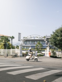 A person wearing a helmet rides a light-colored scooter across a crosswalk on a sunny day. The background features a building with arched structures and palm trees, conveying a relaxed, urban setting.