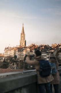 A picturesque view of a European city featuring historic buildings with red-tiled roofs and a prominent, tall church tower. In the foreground, two individuals are seen from behind, one taking a photo with a smartphone. The scene is bathed in warm, late afternoon light, creating a serene and calm atmosphere.