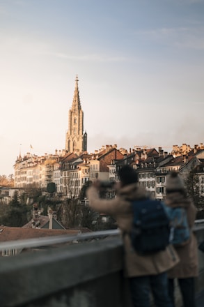 A picturesque view of a European city featuring historic buildings with red-tiled roofs and a prominent, tall church tower. In the foreground, two individuals are seen from behind, one taking a photo with a smartphone. The scene is bathed in warm, late afternoon light, creating a serene and calm atmosphere.