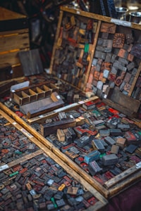 An assortment of vintage wooden letterpress printing blocks laid out in wooden trays and boxes. The blocks display a variety of letters and numbers in different sizes and styles, some of them with visible wear and staining from usage. The scene evokes a sense of history and craftsmanship, capturing the intricate details of a traditional printing method.