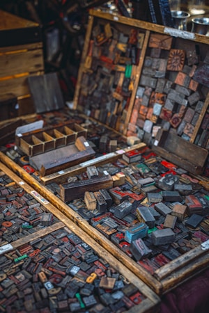 An assortment of vintage wooden letterpress printing blocks laid out in wooden trays and boxes. The blocks display a variety of letters and numbers in different sizes and styles, some of them with visible wear and staining from usage. The scene evokes a sense of history and craftsmanship, capturing the intricate details of a traditional printing method.
