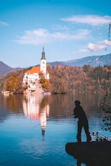 A person is standing on a rock by the water, fishing with a rod. In the background, a picturesque church with an orange roof is situated on a small island in the middle of a serene lake, surrounded by autumn-colored trees and distant mountains. The reflection of the church and trees is visible on the calm water surface.