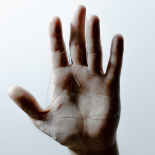 An open hand is centered against a light, neutral background while fine, dark strands are draped across the palm. The intricate pattern formed by the strands contrasts with the smooth skin.
