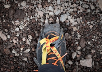A close-up view of a hiking boot with crampons attached, standing on a rocky surface. The laces are orange and yellow, and the boot has visible dirt, indicating use in rugged conditions.