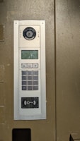 An electronic intercom system mounted on a wall, featuring a camera, a speaker, a numeric keypad, a small display screen, and a sensor for card scanning. The panel appears to be metallic with a sleek design.