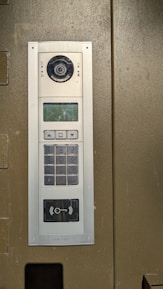 An electronic intercom system mounted on a wall, featuring a camera, a speaker, a numeric keypad, a small display screen, and a sensor for card scanning. The panel appears to be metallic with a sleek design.