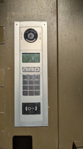 An electronic intercom system mounted on a wall, featuring a camera, a speaker, a numeric keypad, a small display screen, and a sensor for card scanning. The panel appears to be metallic with a sleek design.