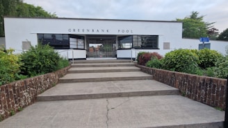 A building with a modern architectural style identified as Greenbank Pool, featuring a white facade with large rectangular windows. There are steps leading up to the entrance, flanked by brick walls. The surrounding area includes well-maintained greenery with various shrubs and plants. A blue sign is visible on the right side, pointing toward another location.