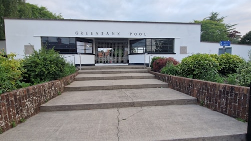 A building with a modern architectural style identified as Greenbank Pool, featuring a white facade with large rectangular windows. There are steps leading up to the entrance, flanked by brick walls. The surrounding area includes well-maintained greenery with various shrubs and plants. A blue sign is visible on the right side, pointing toward another location.
