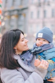 A woman with dark hair is holding a young child dressed in a blue hat and colorful winter jacket. The woman is smiling warmly at the child, and they are both wearing winter clothing, including scarves. The background is softly blurred, showing out-of-focus buildings and winter decorations.