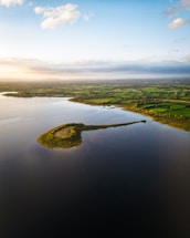 An aerial view of a small, elongated island surrounded by calm waters, with lush, green fields extending into the distance under a clear blue sky. Soft sunlight casts a warm glow on the landscape, adding depth to the variety of greens in the countryside.