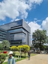 A modern office building with reflective glass windows stands amidst a well-maintained green landscape. The sky is bright with scattered white clouds, and there are a few people walking in the foreground. Manicured bushes and trees surround the area, adding to the serene environment.