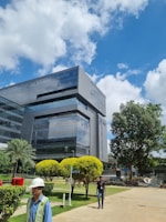 A modern office building with reflective glass windows stands amidst a well-maintained green landscape. The sky is bright with scattered white clouds, and there are a few people walking in the foreground. Manicured bushes and trees surround the area, adding to the serene environment.
