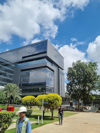 A modern office building with reflective glass windows stands amidst a well-maintained green landscape. The sky is bright with scattered white clouds, and there are a few people walking in the foreground. Manicured bushes and trees surround the area, adding to the serene environment.