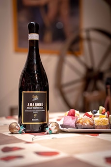A large bottle of Amarone Della Valpolicella wine is placed on a table alongside a plate of assorted colorful pastries. The tablecloth features a red and beige pattern, and festive decorations are set around. In the background, a rustic wooden wheel adds to the cozy ambiance.