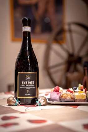 A large bottle of Amarone Della Valpolicella wine is placed on a table alongside a plate of assorted colorful pastries. The tablecloth features a red and beige pattern, and festive decorations are set around. In the background, a rustic wooden wheel adds to the cozy ambiance.