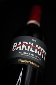 A close-up view of a wine bottle with a label featuring the text 'Bariliot' and 'Aglianico del Vulture.' The label also shows the year 2020 and the word 'Paternoster' in gold font at the bottom. The bottle has a red cap and is set against a dark background, highlighting its elegant presentation.