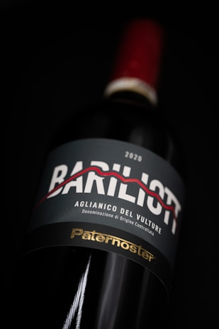 A close-up view of a wine bottle with a label featuring the text 'Bariliot' and 'Aglianico del Vulture.' The label also shows the year 2020 and the word 'Paternoster' in gold font at the bottom. The bottle has a red cap and is set against a dark background, highlighting its elegant presentation.
