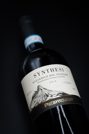 A bottle of wine is displayed against a dark background. The label on the bottle reads 'Synthesi Aglianico del Vulture' and it indicates the year 2018. There is an illustration of a mountain on the label and the word 'Paternoster' is written at the bottom.