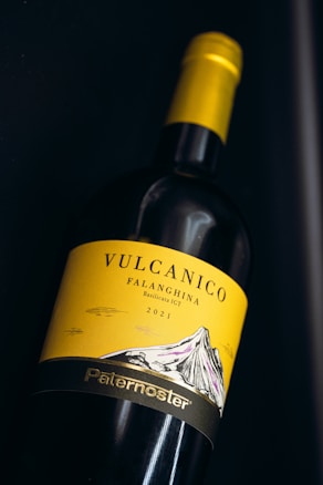 A wine bottle is displayed horizontally, featuring a yellow label with the text 'Vulcanico Falanghina Basilicata IGT 2021' and an illustration of a mountain. The word 'Paternoster' is printed at the bottom of the label in a metallic finish. The bottle has a yellow cap, and the background is dark.