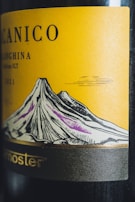 A close-up view of a wine bottle label featuring an illustrated mountain with distinctively defined lines and shades. The background of the label is yellow, and the illustration includes gray and purple hues for the mountain. Text on the label is partially visible at the top and bottom.
