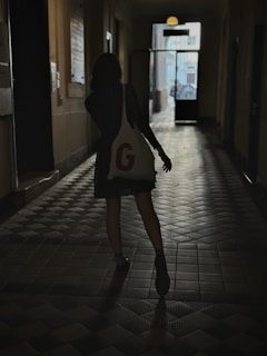 A silhouette of a person walking down a dimly lit corridor, carrying a bag with a large letter 'G' on it. The floor has a tiled pattern and the far end of the corridor is illuminated by natural light coming through an open door, revealing the outdoor area.