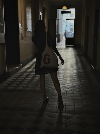 A silhouette of a person walking down a dimly lit corridor, carrying a bag with a large letter 'G' on it. The floor has a tiled pattern and the far end of the corridor is illuminated by natural light coming through an open door, revealing the outdoor area.