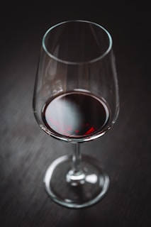 A glass of red wine sits on a dark, textured surface. The wine contrasts with the clear glass and has a rich, deep red color that reflects some light.