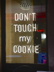 A neon sign displays the phrase 'DON'T TOUCH MY COOKIE' in bright white light against a dark background. Above the text is an image of a cookie with heart-shaped patterns. Reflections and shadows are visible, suggesting the sign is inside a glass window.