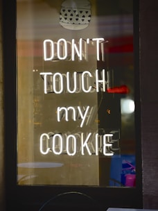 A neon sign displays the phrase 'DON'T TOUCH MY COOKIE' in bright white light against a dark background. Above the text is an image of a cookie with heart-shaped patterns. Reflections and shadows are visible, suggesting the sign is inside a glass window.