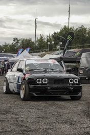 A modified race car with bold customizations, featuring black and white body detailing and headlights resembling rings. The car is parked on a gravel surface at an automotive event, surrounded by large branded tents and banners. There are a few individuals in the background near the tents.