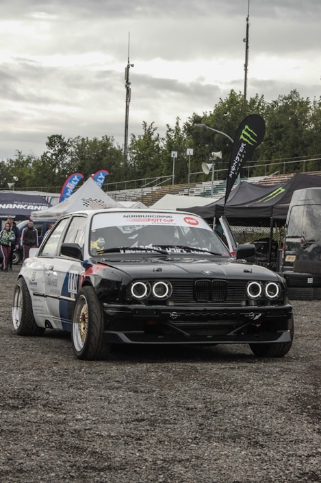 A modified race car with bold customizations, featuring black and white body detailing and headlights resembling rings. The car is parked on a gravel surface at an automotive event, surrounded by large branded tents and banners. There are a few individuals in the background near the tents.