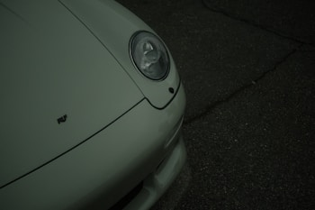 A close-up view of a light-colored sports car's front end, focusing on the headlight and hood. The car is parked on an asphalt surface, creating a sleek and polished appearance.