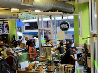A busy cafe environment with several staff and customers interacting near the counter. The decor features green walls and a prominent duct system. There are pastries displayed in a glass case, and a menu board is visible. Outside, a bus is passing by the window.
