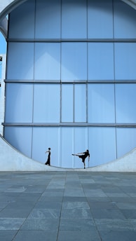 Two individuals dressed in black, positioned within a large circular cut-out of a modern architectural structure featuring light blue panels. One person is standing straight, while the other is performing a high kick. The ground is paved with gray tiles.