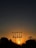 A silhouetted power line tower stands against a vibrant sunset sky, with the sun glowing brightly near the horizon. The scene features trees in the foreground, which are also silhouetted, adding depth to the composition.