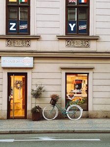 A quaint storefront with beige walls featuring decorative stonework. Two windows above bear educational signage and various national flags. Below, a welcoming doorway is adorned with a floral wreath. Beside the entrance, a teal vintage bicycle with a basket containing dried flowers leans against the wall. A potted plant enhances the homely feel, and the shop sign reads 'MINICHLEBICKY Ema.'