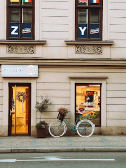 A quaint storefront with beige walls featuring decorative stonework. Two windows above bear educational signage and various national flags. Below, a welcoming doorway is adorned with a floral wreath. Beside the entrance, a teal vintage bicycle with a basket containing dried flowers leans against the wall. A potted plant enhances the homely feel, and the shop sign reads 'MINICHLEBICKY Ema.'