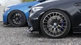 Close-up of two cars parked side by side, focusing on their wheels. The car on the left is blue, while the car on the right is black. Both vehicles have sporty, intricate alloy wheel rims and performance brake discs with calipers visible.