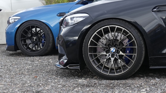 Close-up of two cars parked side by side, focusing on their wheels. The car on the left is blue, while the car on the right is black. Both vehicles have sporty, intricate alloy wheel rims and performance brake discs with calipers visible.