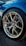 A close-up of a performance car's wheel, featuring a detailed view of the alloy wheel design with a prominent BMW logo at the center. The tire is from Michelin, and the brake calipers and rotors are visible behind the wheel. The car has a shiny blue exterior, reflecting the surrounding environment.