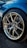 A close-up of a performance car's wheel, featuring a detailed view of the alloy wheel design with a prominent BMW logo at the center. The tire is from Michelin, and the brake calipers and rotors are visible behind the wheel. The car has a shiny blue exterior, reflecting the surrounding environment.