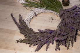 A partially made circular wreath lies on a light wooden surface. The wreath is adorned with numerous small purple flowers, presumably lavender, attached to a base of green stems. There is also a small spool of thread or twine nearby, suggesting ongoing or recent crafting activity.