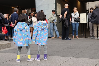 Two children wearing colorful butterfly-patterned blue jackets and matching leggings stand on a paved area. They are facing a group of adults who appear to be waiting, some casually standing around, while others are looking in different directions. The scene takes place outdoors, possibly near an entrance or exit as indicated by a sign in the background.